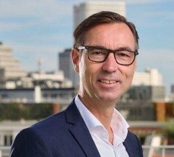 Mann im Anzug mit Brille, lächelnd vor urbaner Kulisse mit Hochhäusern im Hintergrund.