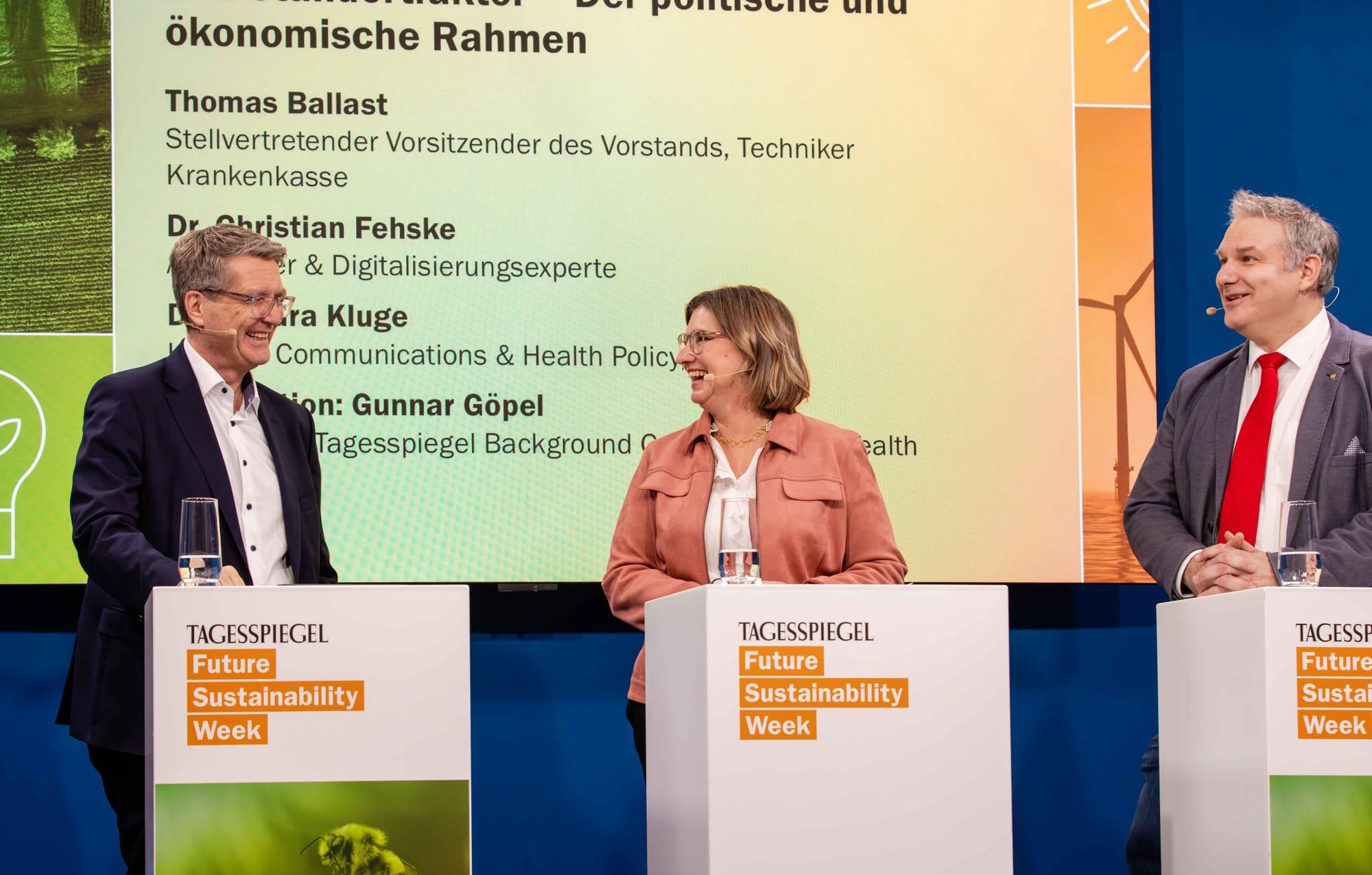 Drei Personen stehen an Pulten vor einer Leinwand mit Text zu ökonomischen Rahmen und Gesundheit.