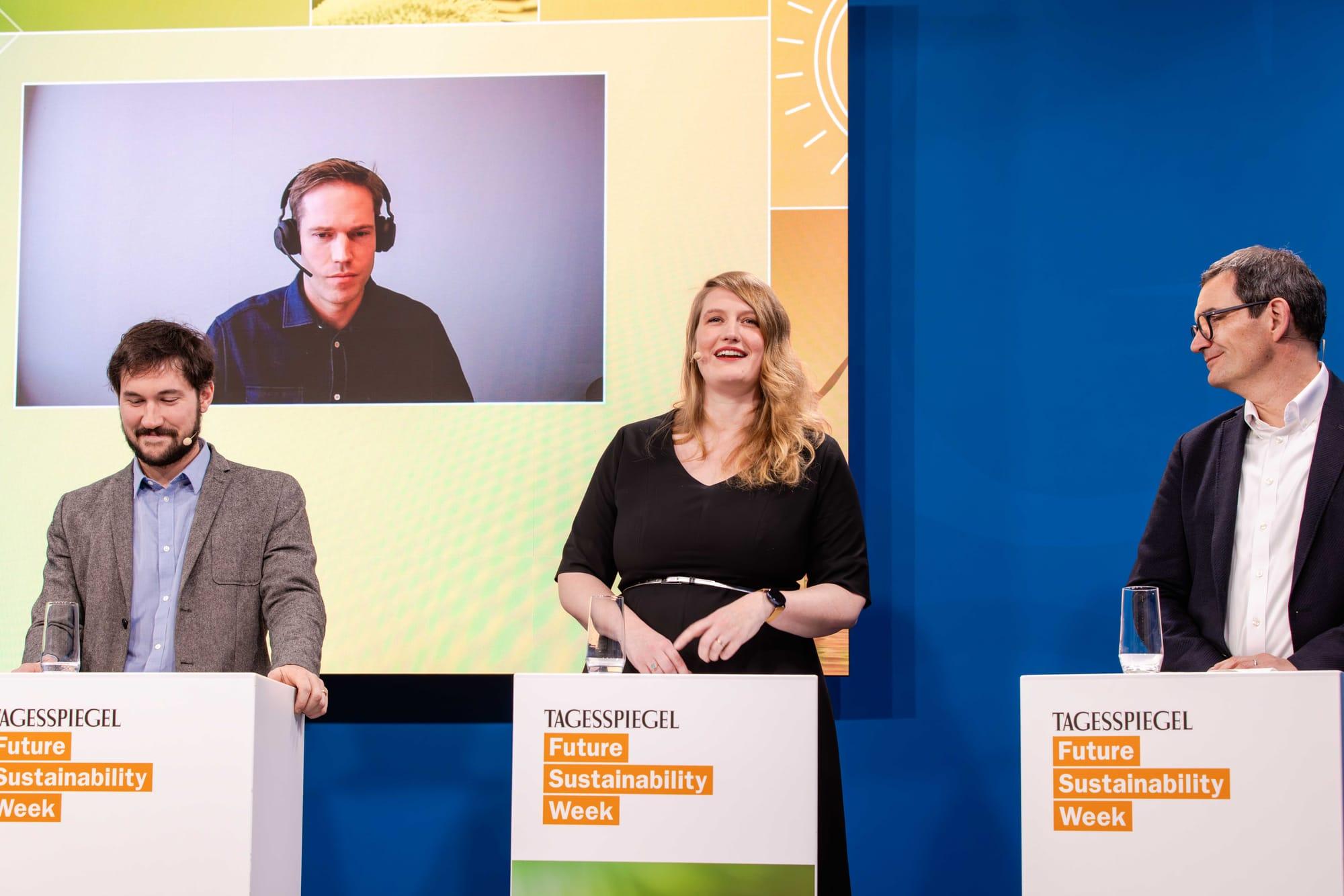 Diskussion auf der Bühne mit drei Personen und einer zugeschalteten Person auf dem Bildschirm