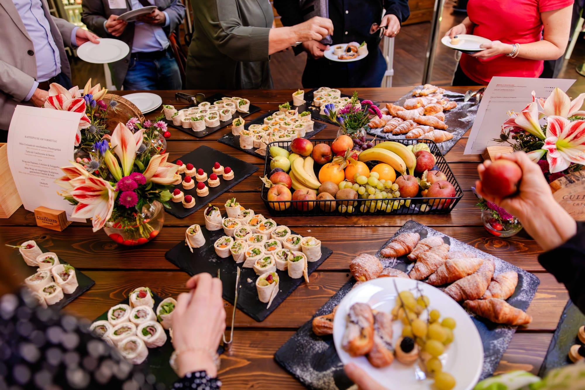 Buffet mit Obst, Croissants, Wraps und kleinen Häppchen, dekoriert mit Blumen auf einem Holztisch