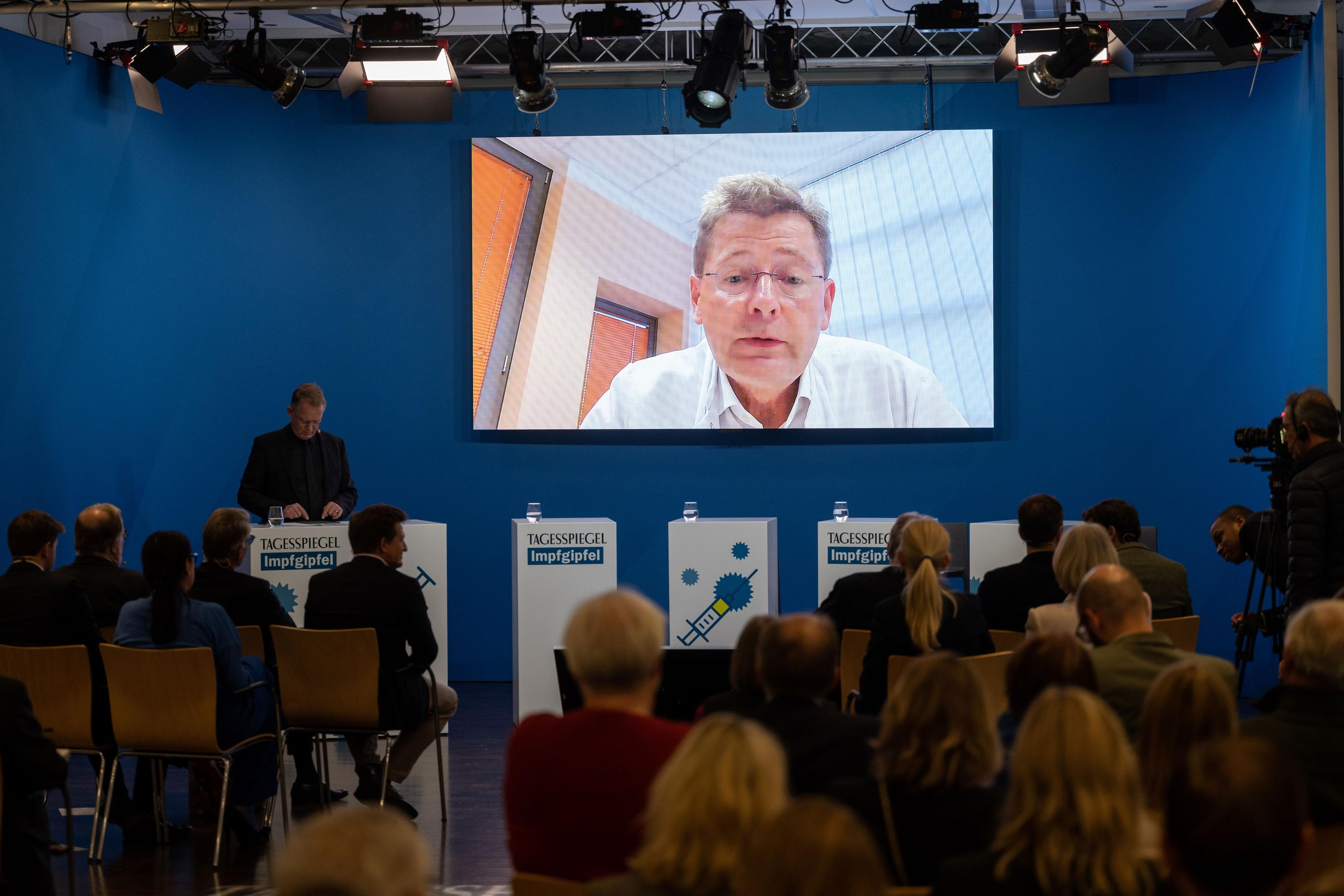 Ein Mann steht vor Publikum hinter einem Rednerpult. Auf einem Screen ist eine Person dazugeschaltet
