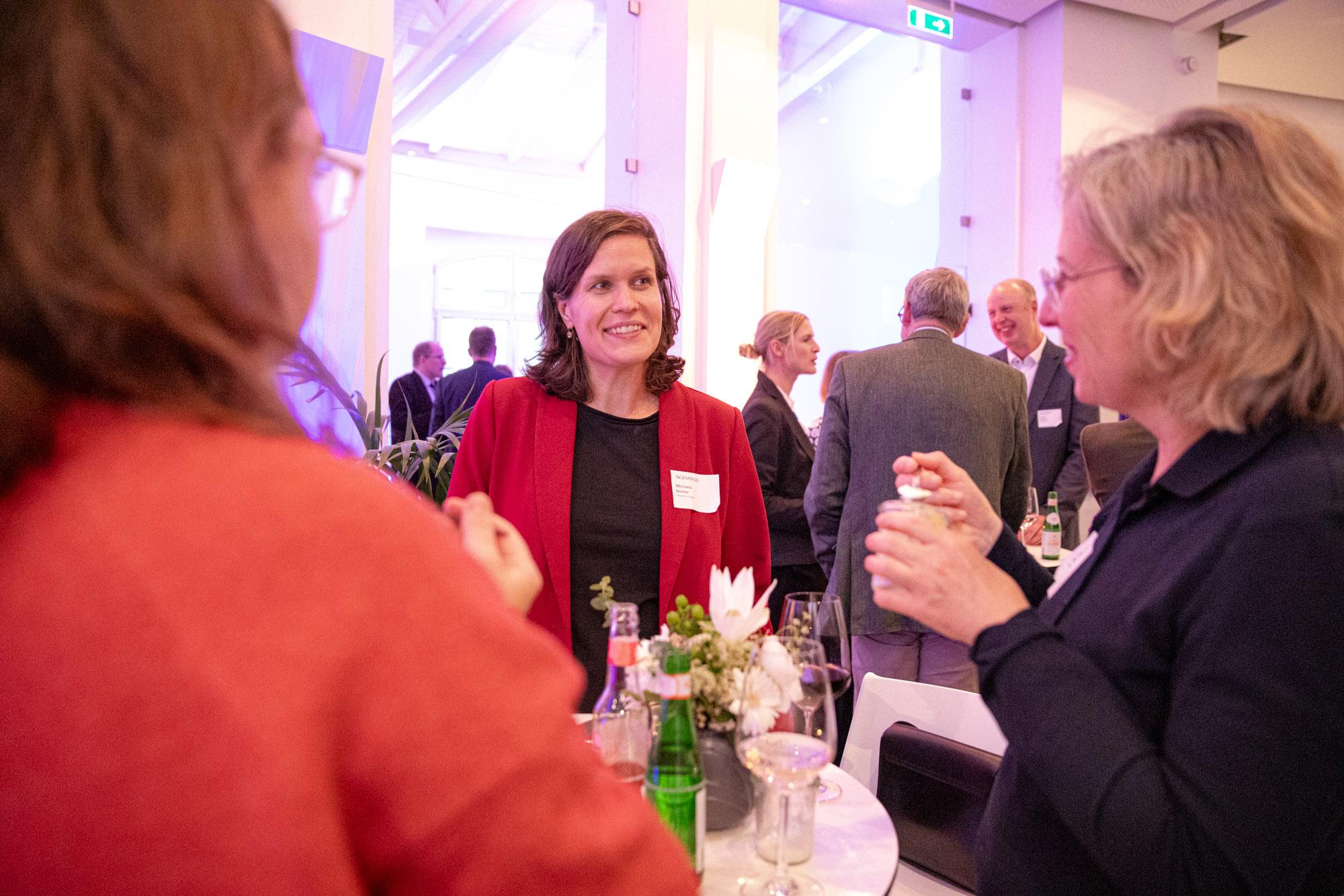 Drei Frauen im Gespräch an einem Stehtisch, die mittlere trägt einen roten Blazer.