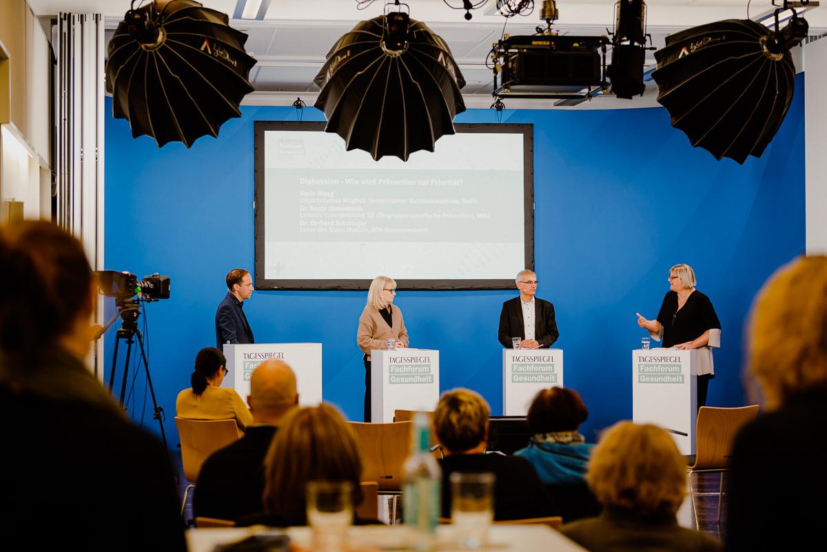 Vor Publikum stehen vier Personen jeweils hinter einem Rednerpult. Im Hintergrund ein Screen.