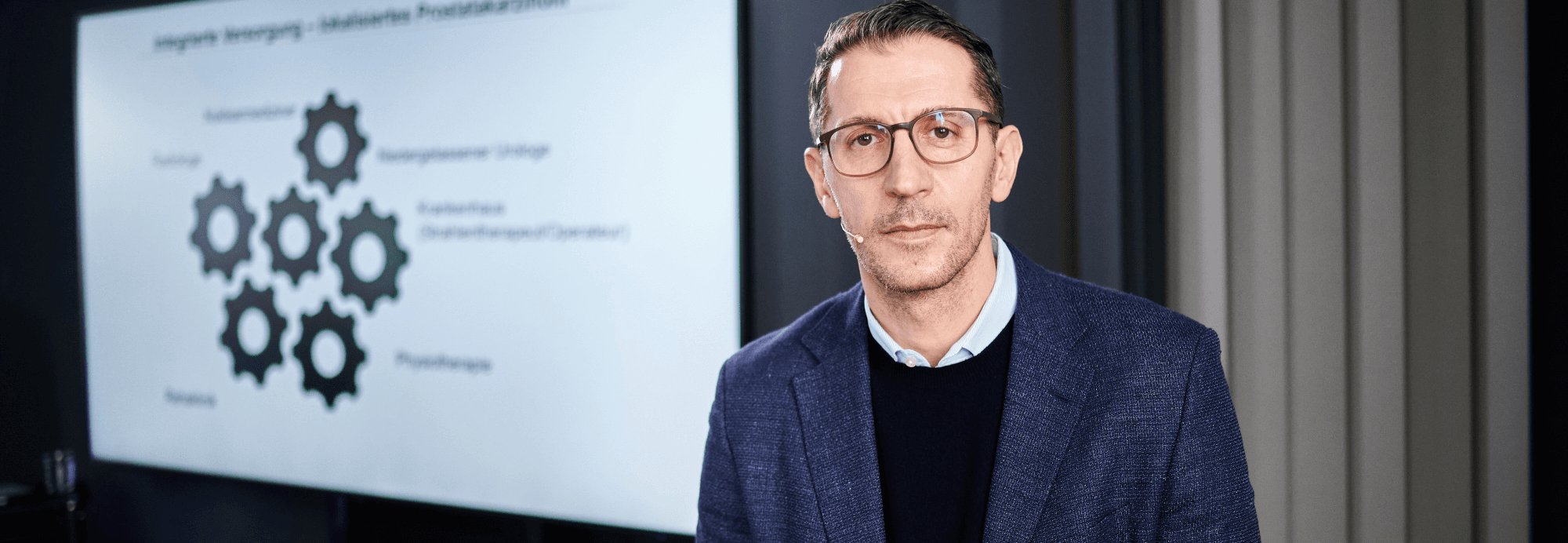 Ein Mann mit Brille im Anzug steht vor einer Leinwand. Auf der Leinwand sind Zahnräder.