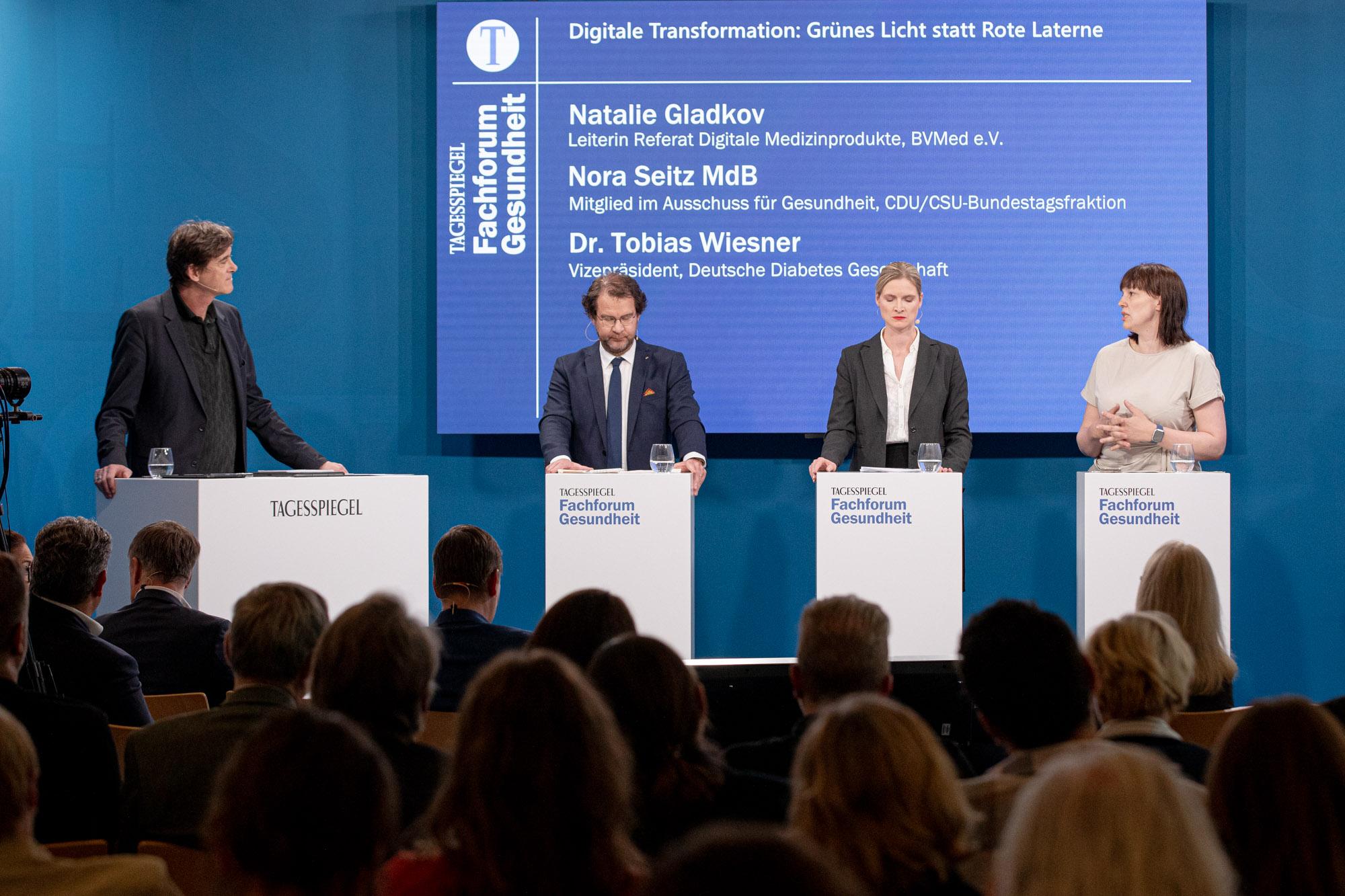 Vier Personen an Pulten bei Podiumsdiskussion vor blauem Bildschirm, Publikum davor.