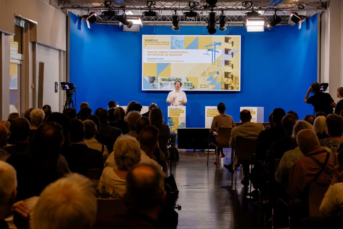 Eine Person spricht am Pult vor Publikum, im Hintergrund ein Screen.