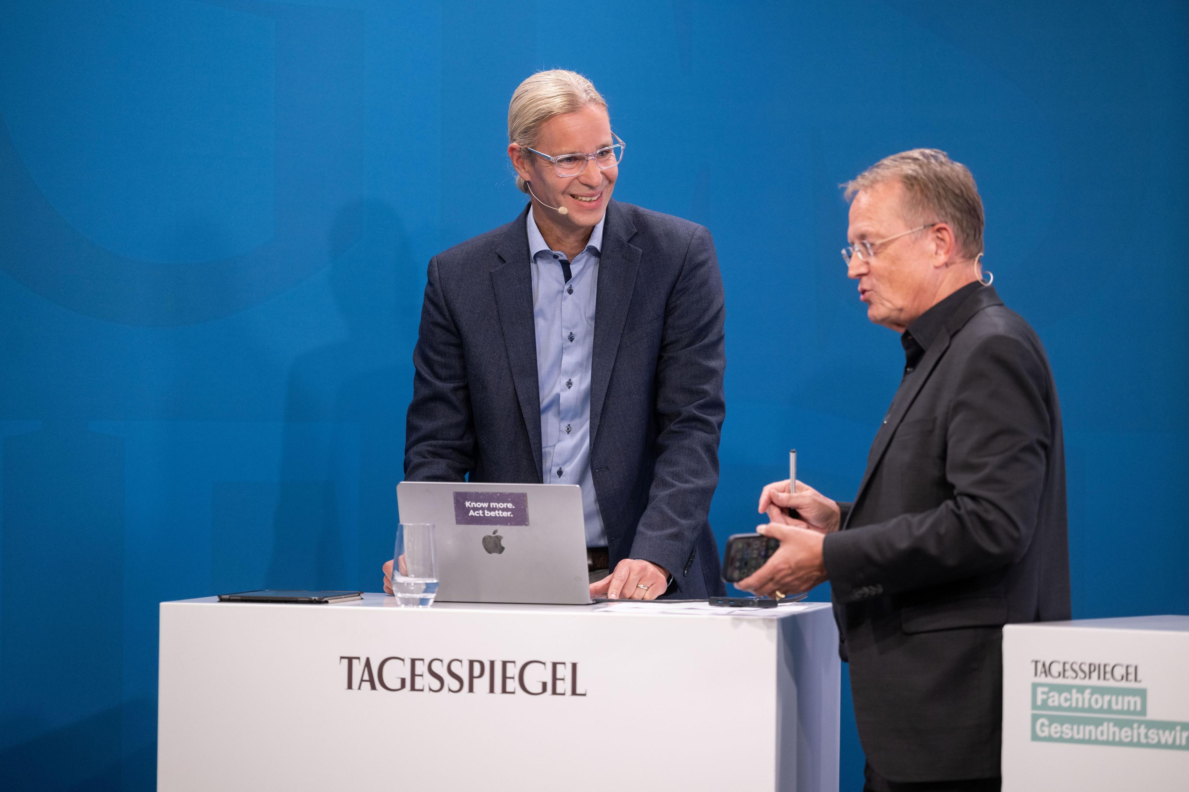 Zwei Personen am „Tagesspiegel“-Podium, eine spricht, die andere nutzt Laptop, blauer Hintergrund.