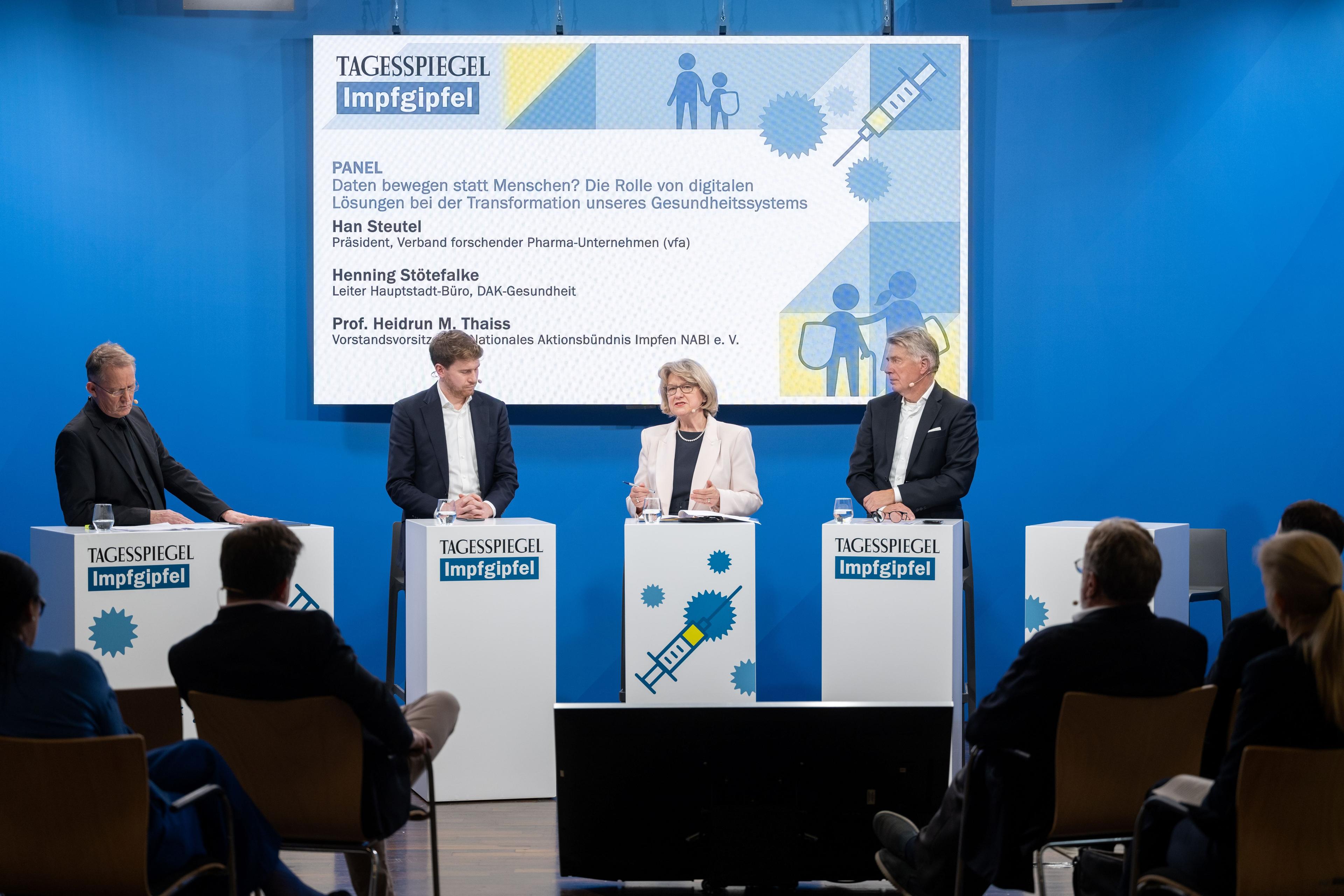 Vier Personen diskutieren auf Bühne beim Impfgipfel, Bildschirm im Hintergrund.
