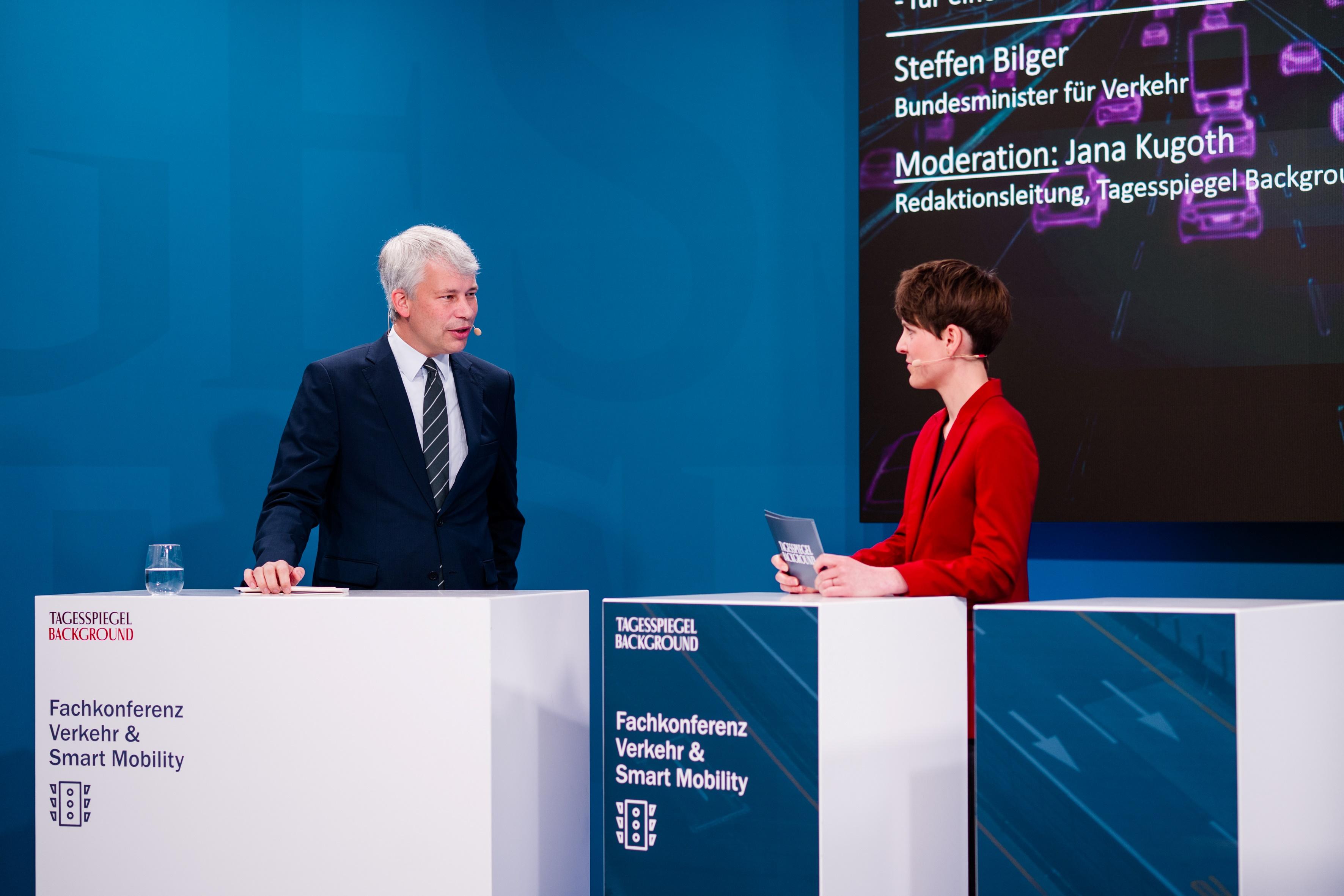 Ein Redner und die Moderatorin stehen auf der Bühne und sprechen miteinander.