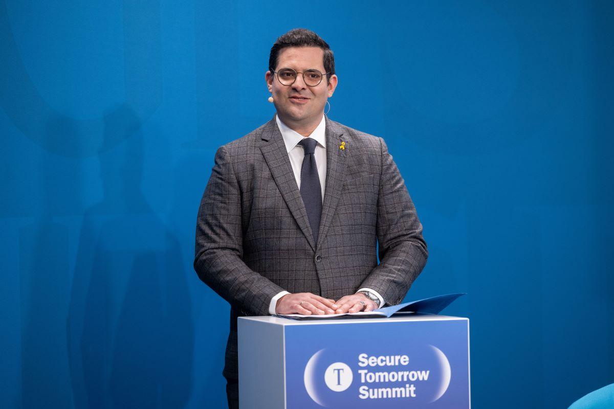 Sassan Parvin am Pult „Secure Tomorrow Summit“ auf blauer Bühne, Ordner auf dem Pult.