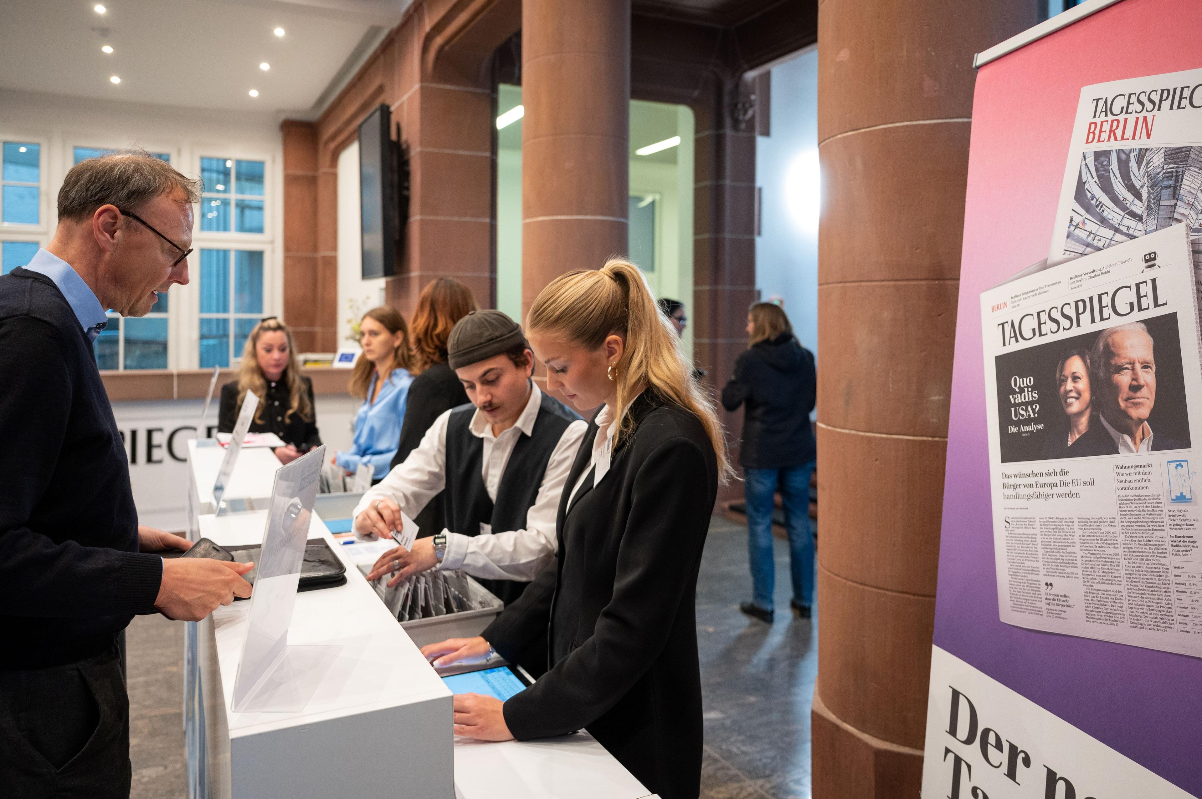 Menschen am Check-in-Schalter eines Events, rechts ein großes Tagesspiegel-Poster.