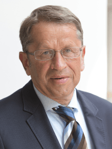 Ein Mann, mit kurzen Haaren, der eine Brille und ein Anzug trägt.