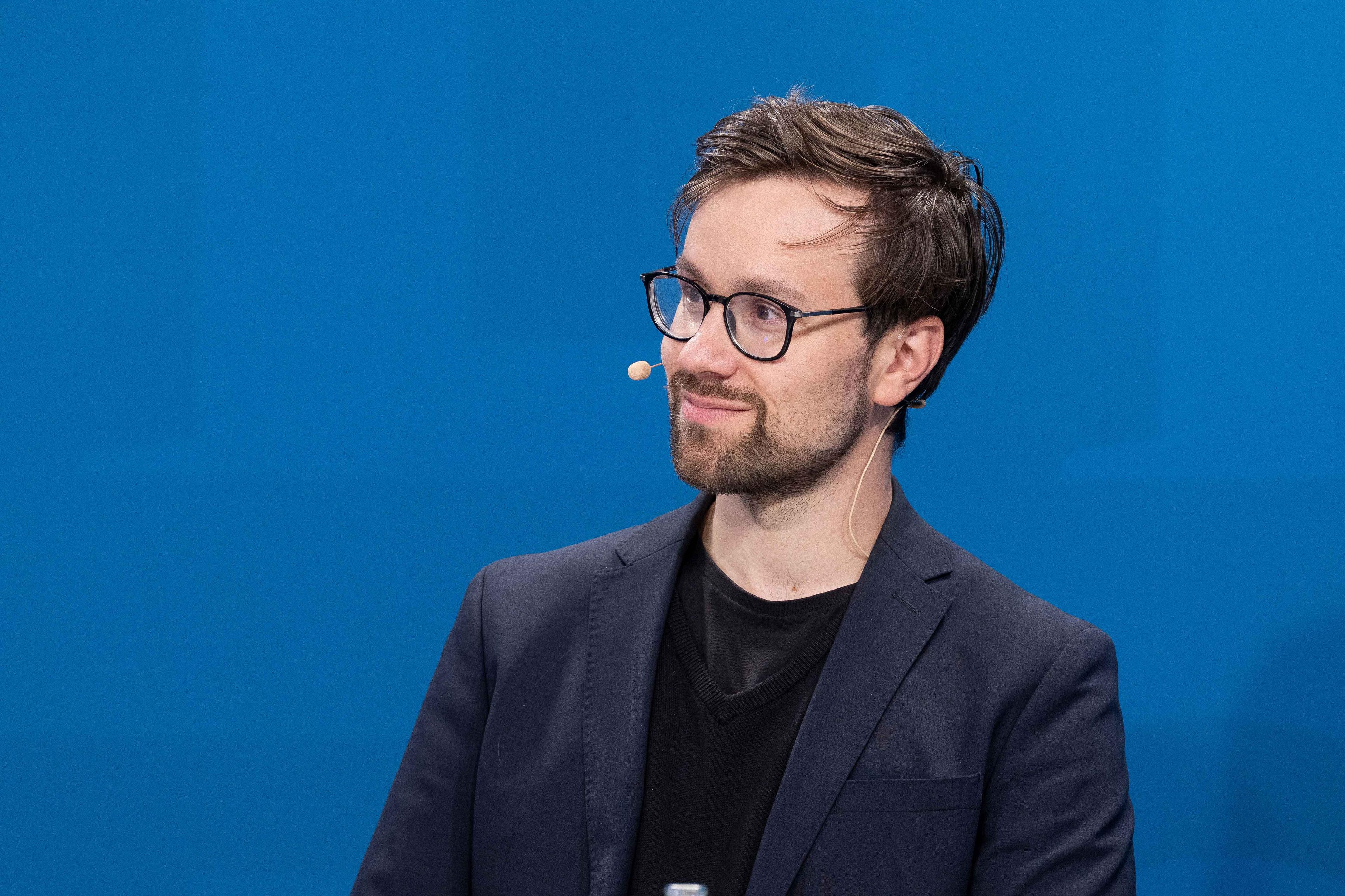 Porträt von Johannes Wagner mit Brille und Headset-Mikrofon vor blauem Hintergrund.