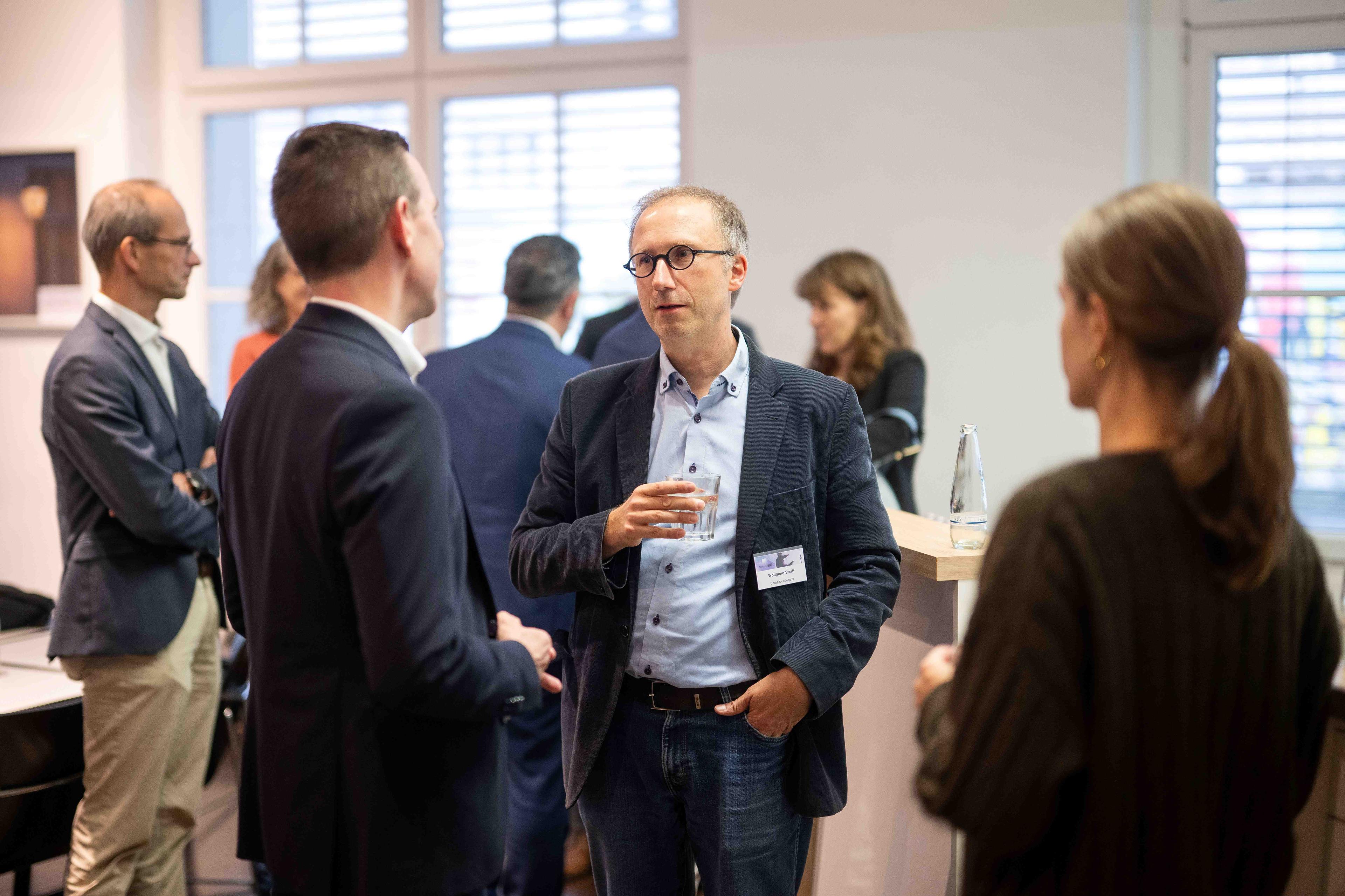 Ein Mann hält ein Wasserglas und unterhält sich mit Gästen beim Fachforum.