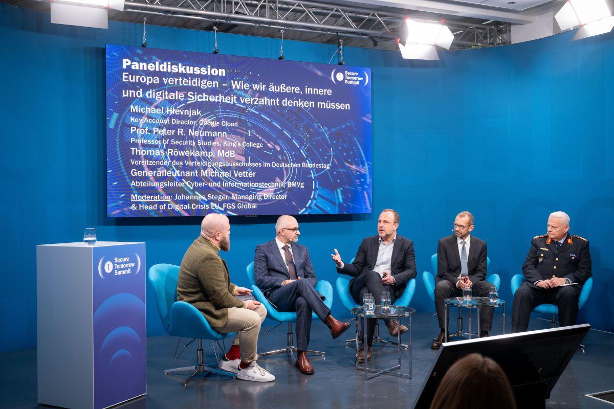 Panel mit fünf Personen auf blauer Bühne, große LED‑Tafel „Europa verteidigen“.