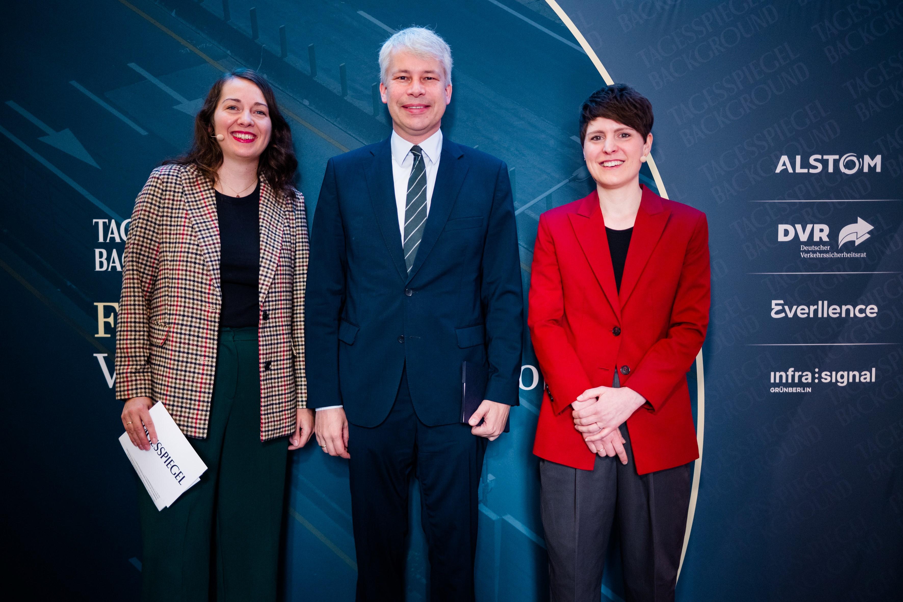 Drei lächelnde Personen posieren vor der gebrandeten Kulisse der Fachkonferenz Verkehr & Smart Mobility.