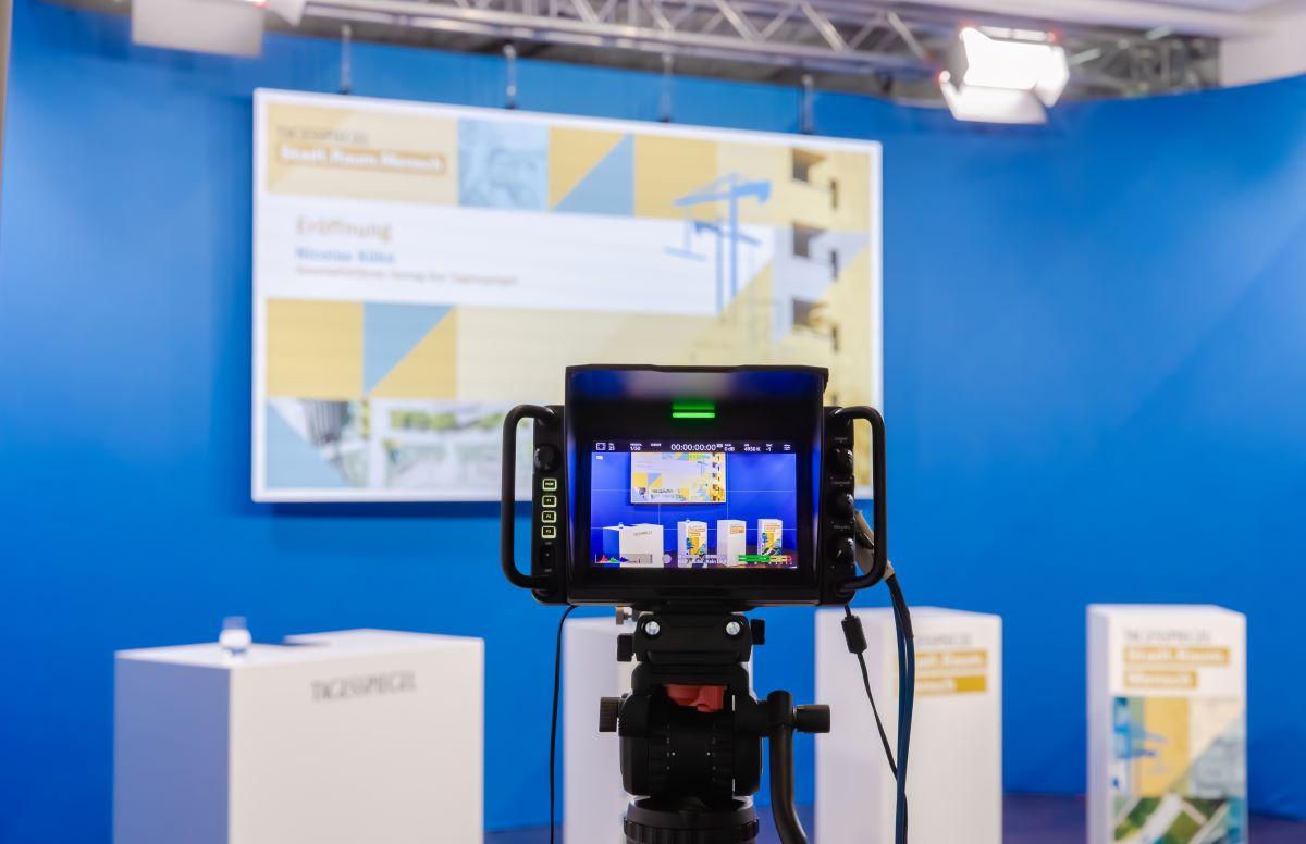 Eine Kamera filmt drei Rednerpulte vor blauem Hintergrund mit Präsentationsscreen.