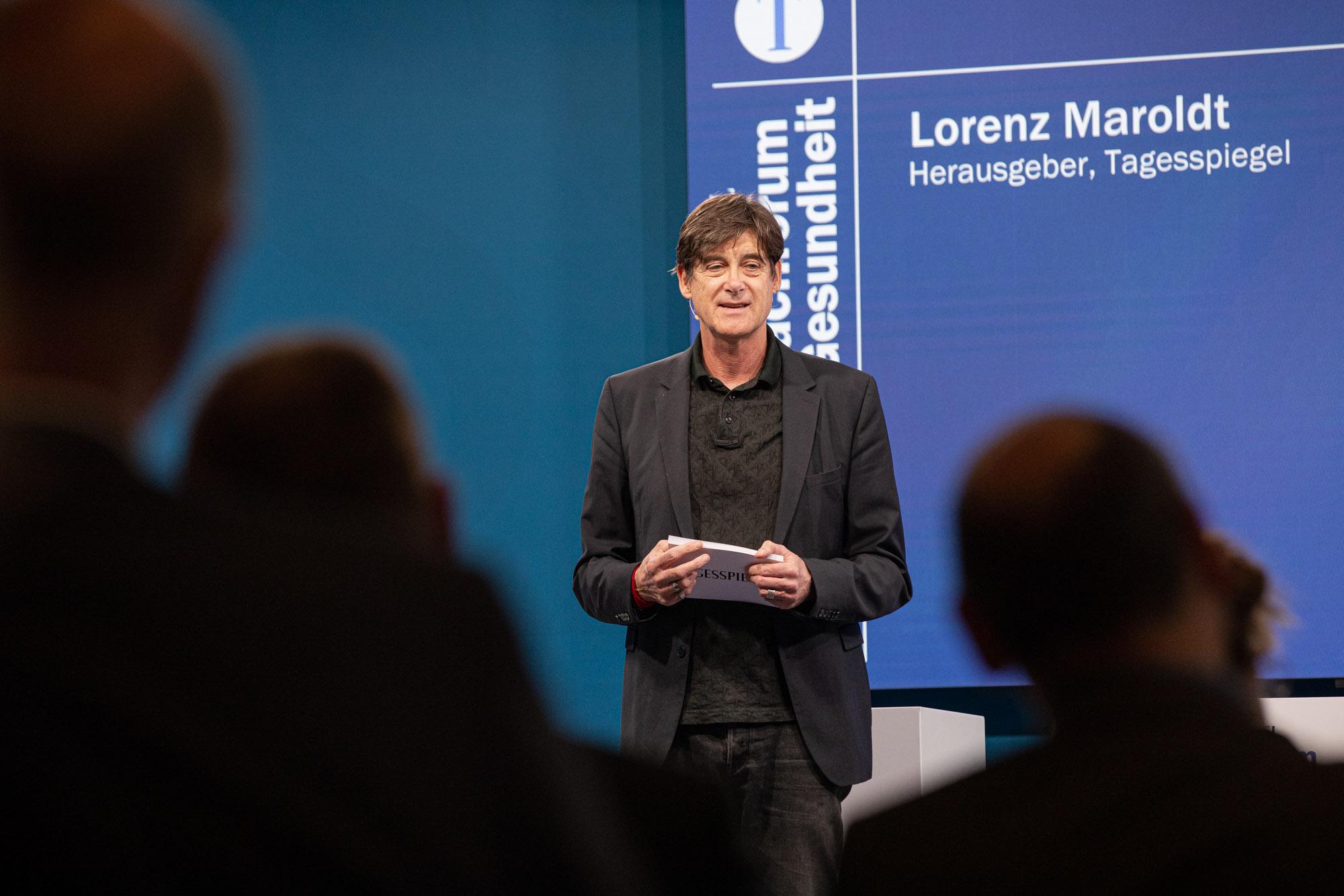 Lorenz Maroldt spricht beim Tagesspiegel Fachforum Gesundheit vor blauem Infobildschirm.
