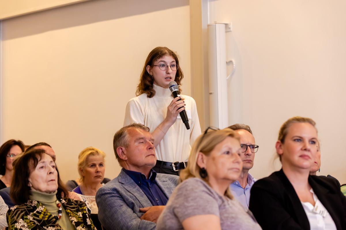 Im Publikum steht eine Frau mit Mikrofon.