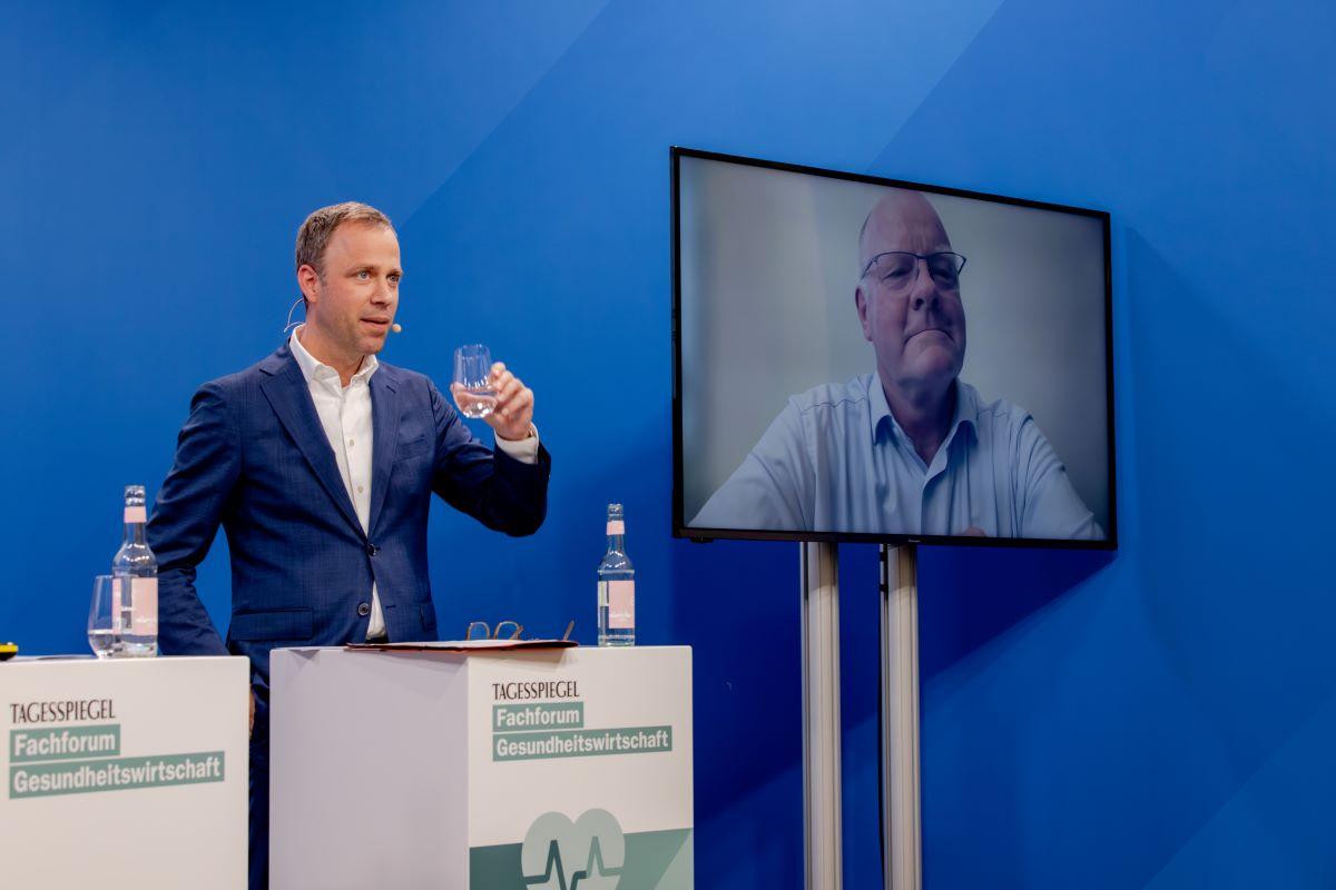 Ein Mann hinter einem Rednerpult. Neben ihm ist ein Mann virtuell zugeschaltet.