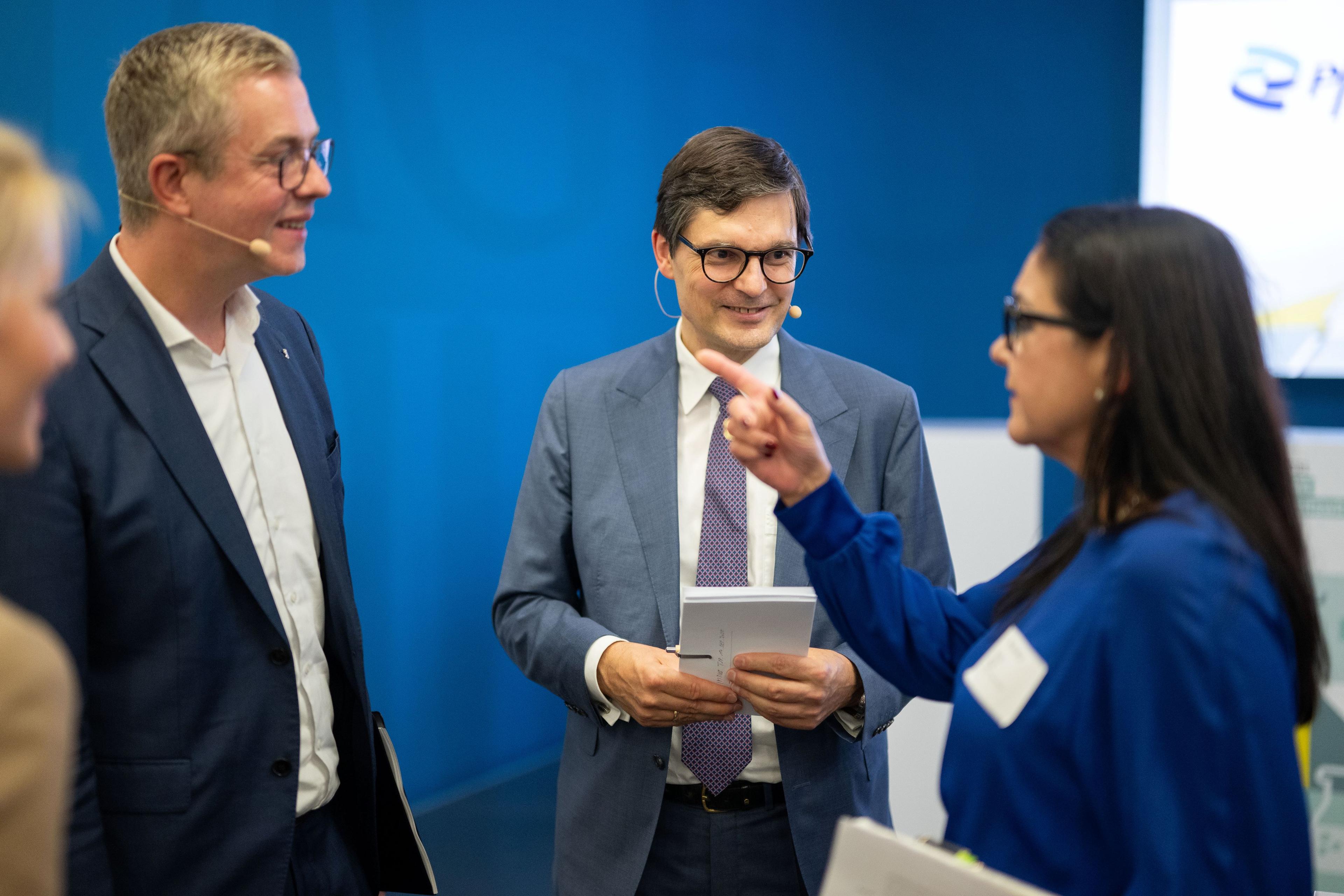 Vier Personen im Gespräch bei Konferenz, blaue Kulisse, zwei mit Notizblöcken, eine Person zeigt.