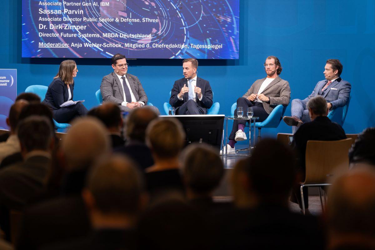Panelrunde auf blauer Bühne; Moderatorin links, vier Gäste, Publikum unscharf im Vordergrund.