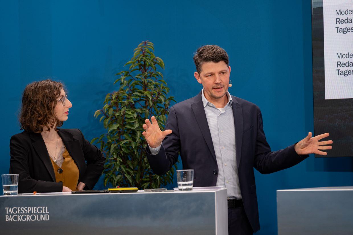 Zwei Personen am Pult im Gespräch vor blauer Wand.