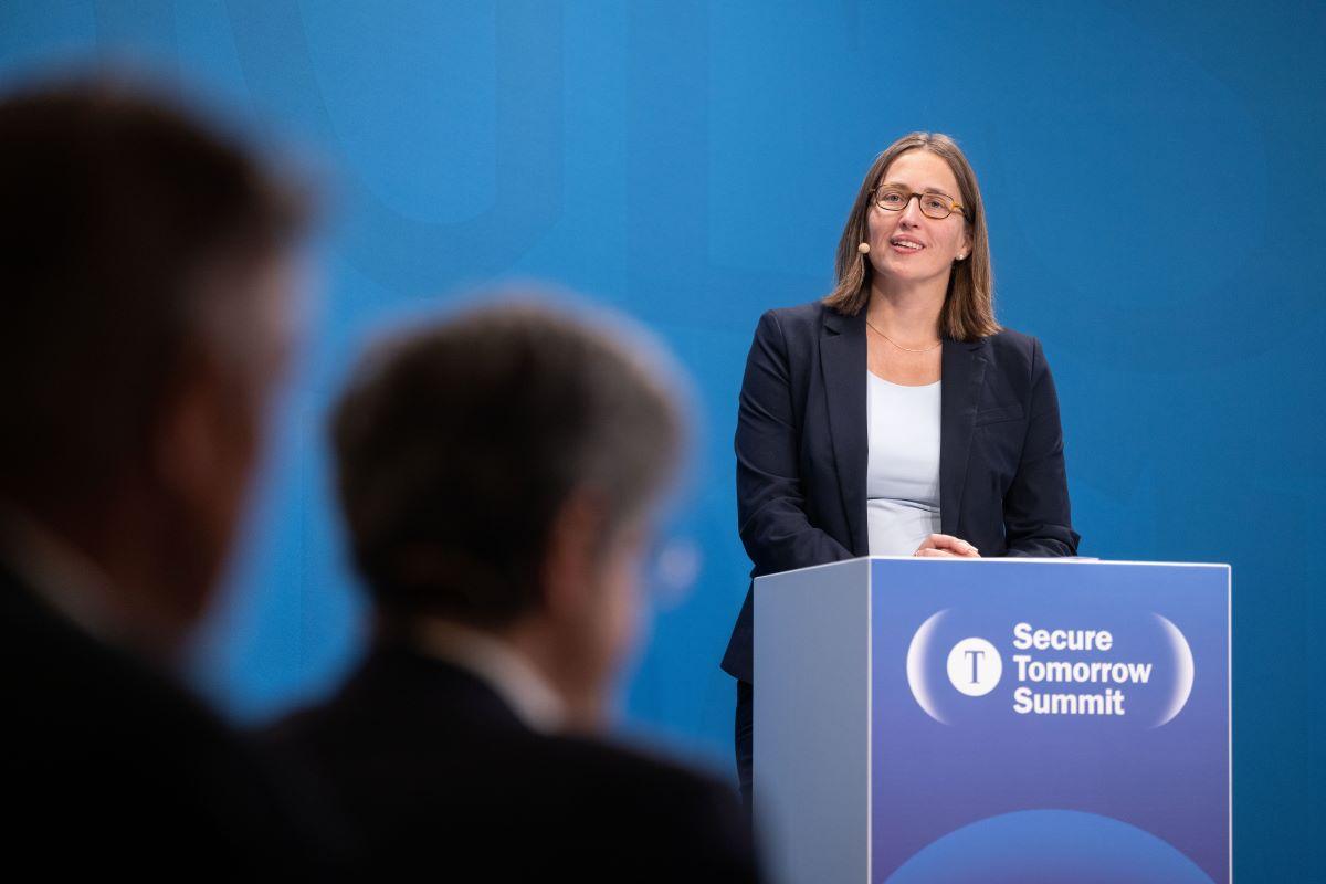 Anja Wehler-Schöck am Pult mit Veranstaltungslogo, blaue Bühne; Publikum unscharf im Vordergrund.
