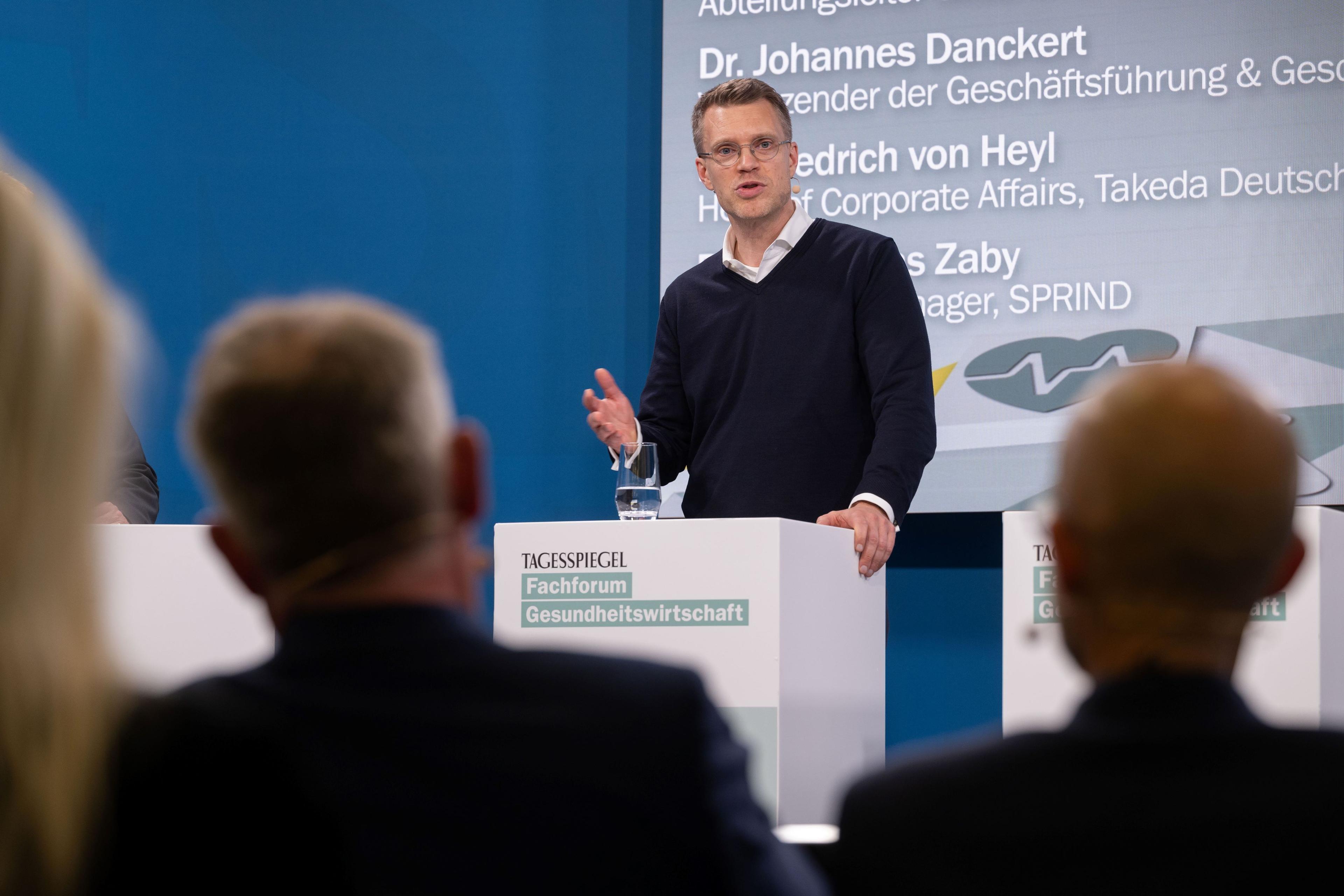 Person spricht am „Tagesspiegel“-Podium, drei Zuhörer im Vordergrund, Namen im Hintergrund.