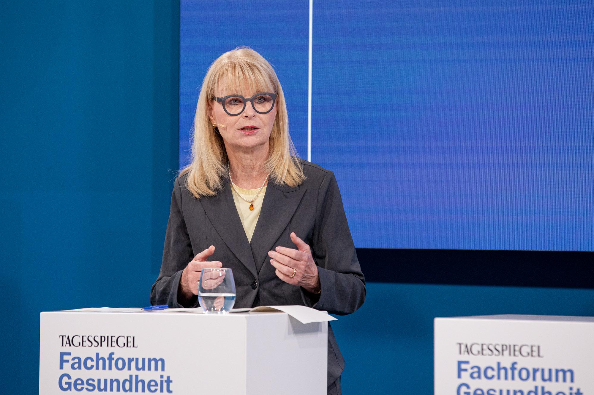 Blonde Frau mit Brille und dunklem Blazer spricht an weißem Pult auf Fachforum Gesundheit.