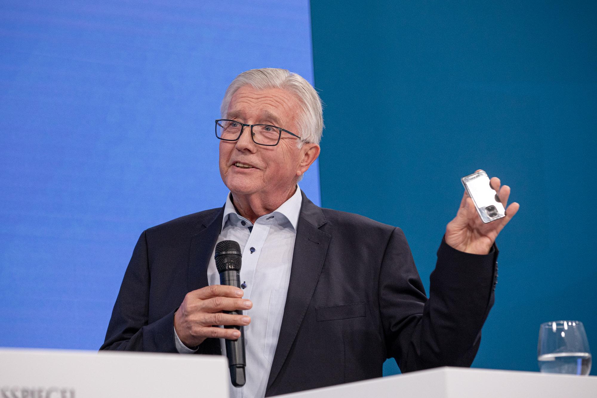 Älterer Mann mit Brille hält Mikrofon und kleines silbernes Gerät beim Fachforum Gesundheit.