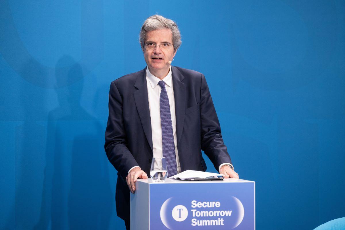 François Delattre am Pult „Secure Tomorrow Summit“ auf blauer Bühne, Glas Wasser daneben.
