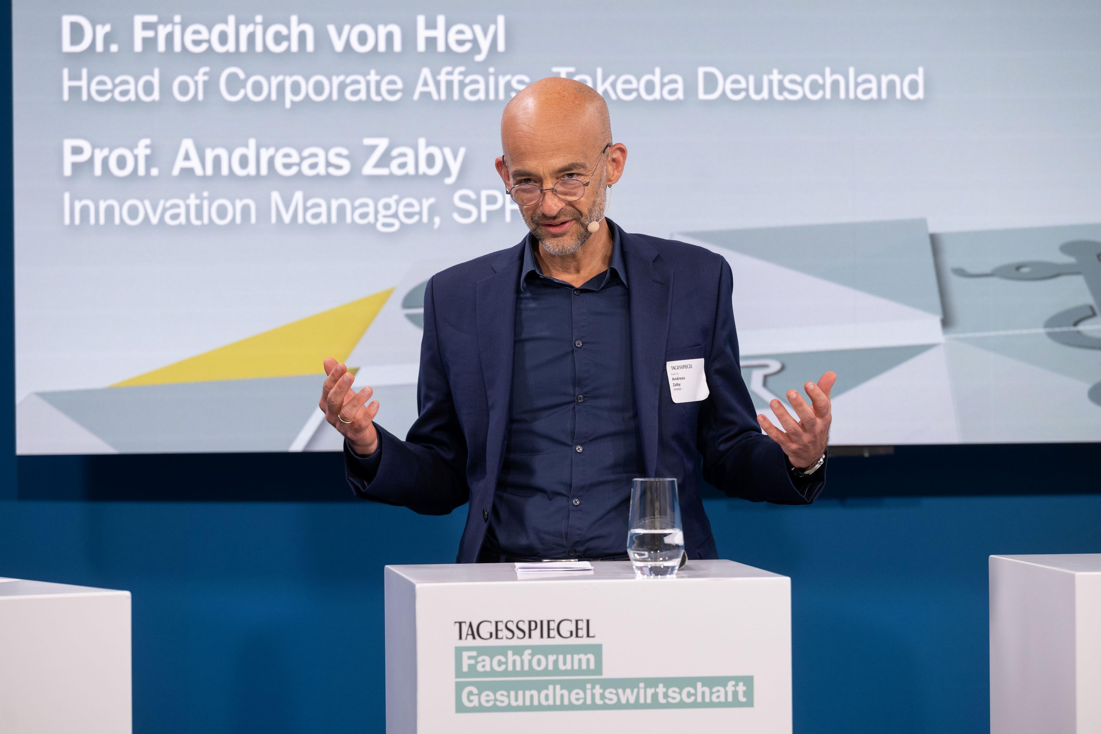 Person spricht am „Tagesspiegel“-Podium, Hintergrund zeigt Namen von zwei Experten.