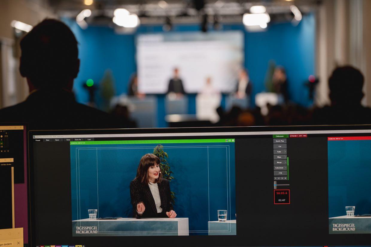 Monitor zeigt Live‑Produktion, Podium im Hintergrund.