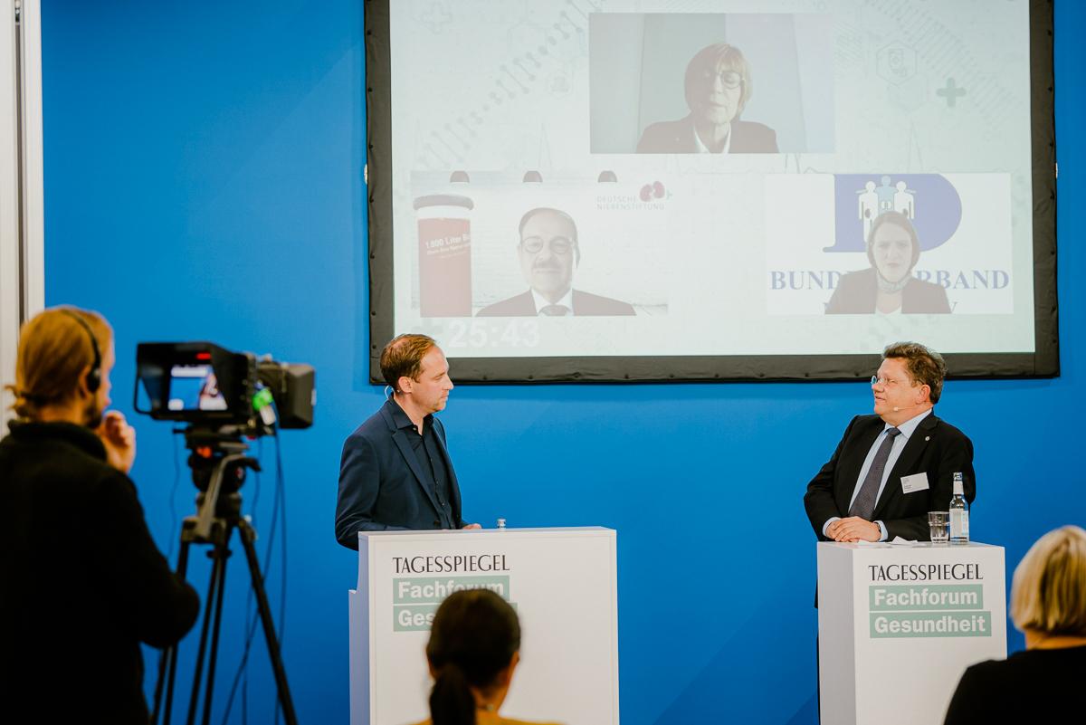 Personen stehen jeweils hinter einem Rednerpult. Auf Screen sind drei Personen virtuell zugeschaltet