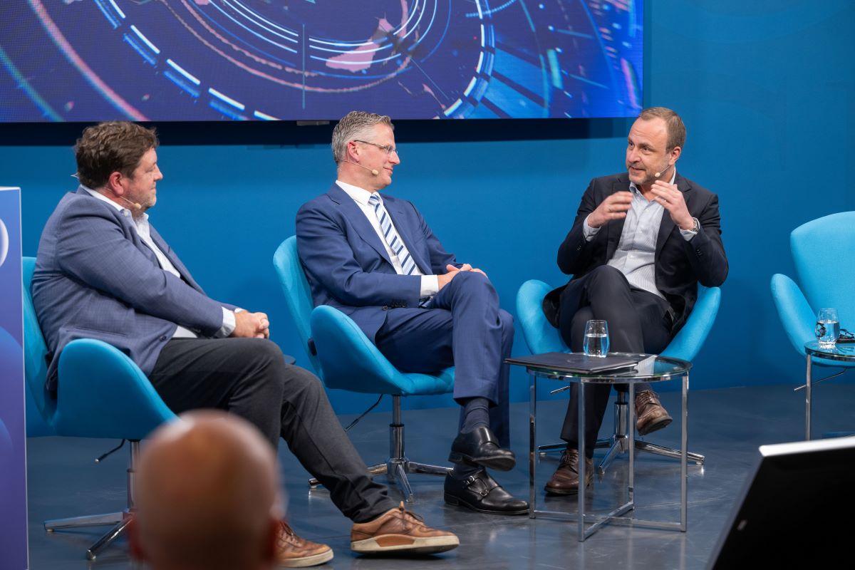 Drei Panelisten im Gespräch auf blauer Bühne, Gläser auf kleinen Tischen.