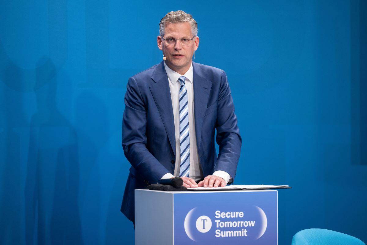 Christoph de Vries am Pult mit Notizen, „Secure Tomorrow Summit“-Logo, blaue Rückwand.