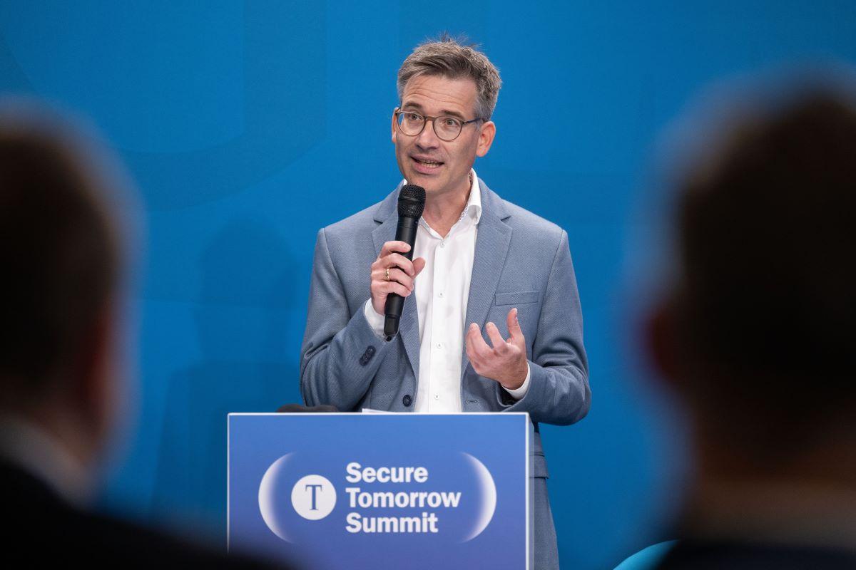 Christian Tretbar mit Mikrofon am Pult „Secure Tomorrow Summit“, Publikum im Vordergrund.