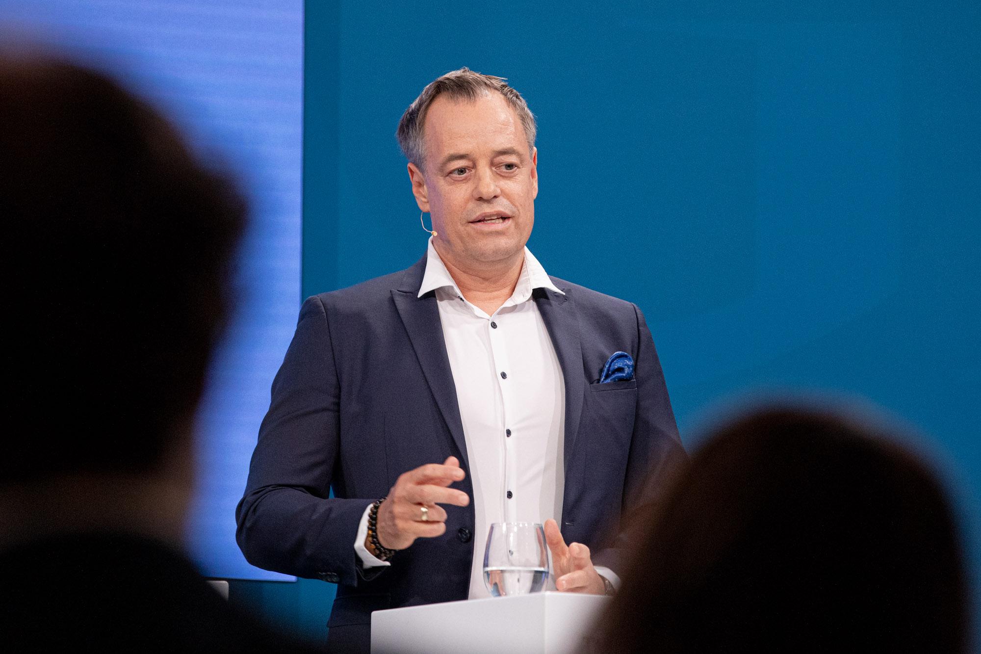 Mann im Anzug spricht gestikulierend am Pult beim Fachforum Gesundheit vor blauem Hintergrund.