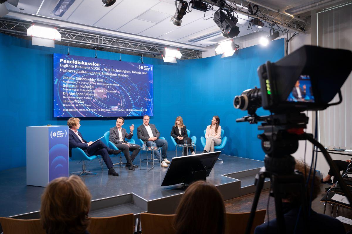 Kamera filmt Panel „Digitale Resilienz 2030“; fünf Gäste auf blauer Bühne, LED‑Tafel im Hintergrund.