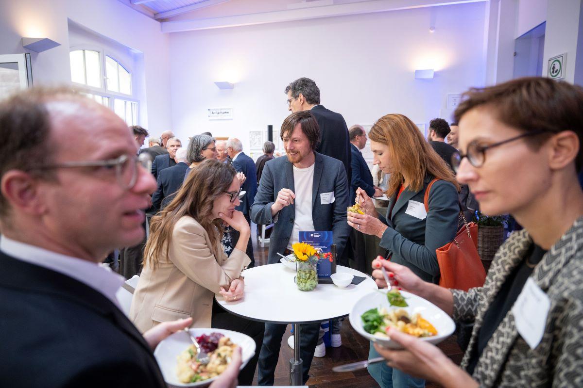 Foyer voll mit Gästen beim Networking; helle Fenster, Stehtische, Gespräche, Essen.
