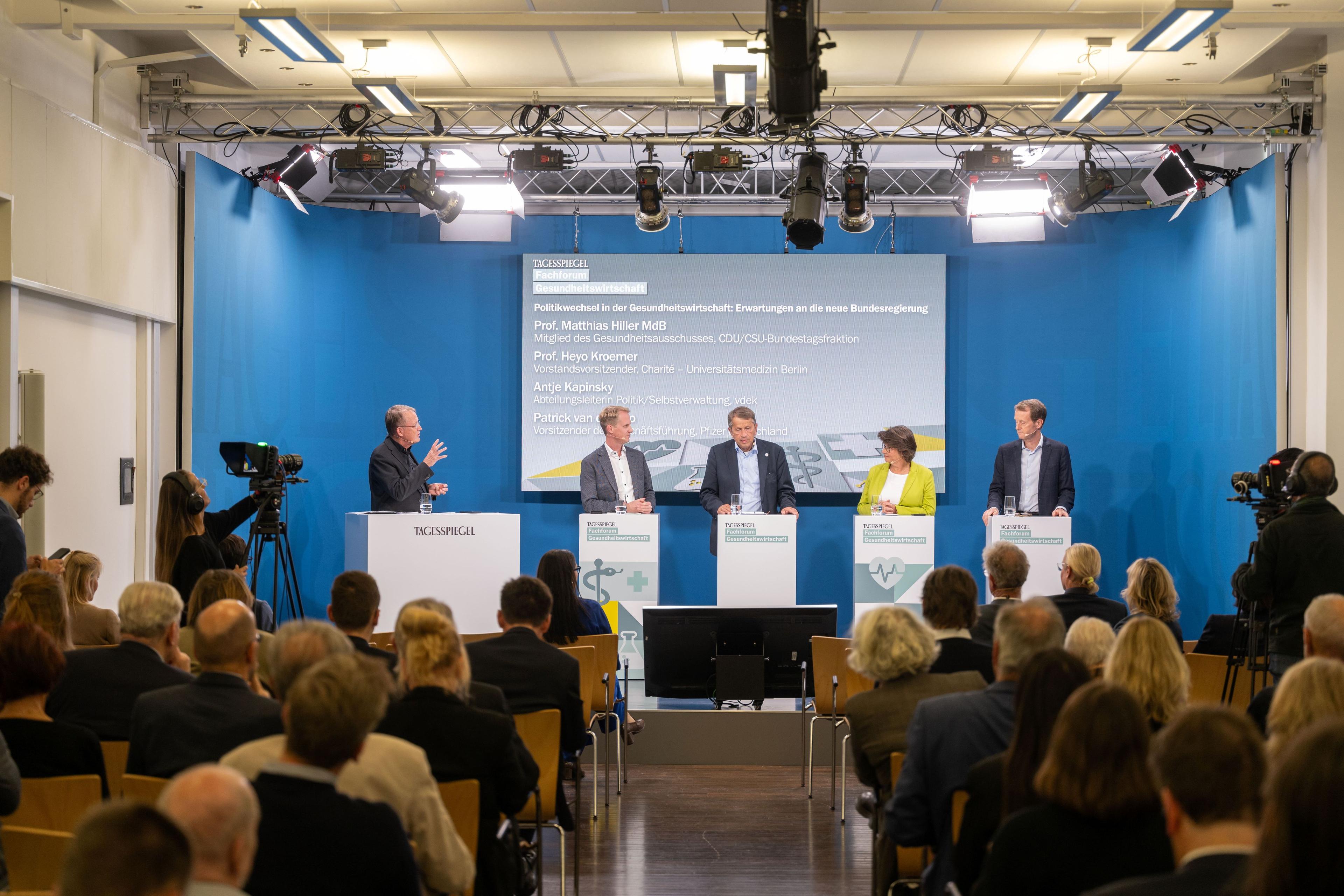 Fünf Vortragende auf Bühne bei öffentlicher Vorlesung, Publikum und blaue Präsentationswand.