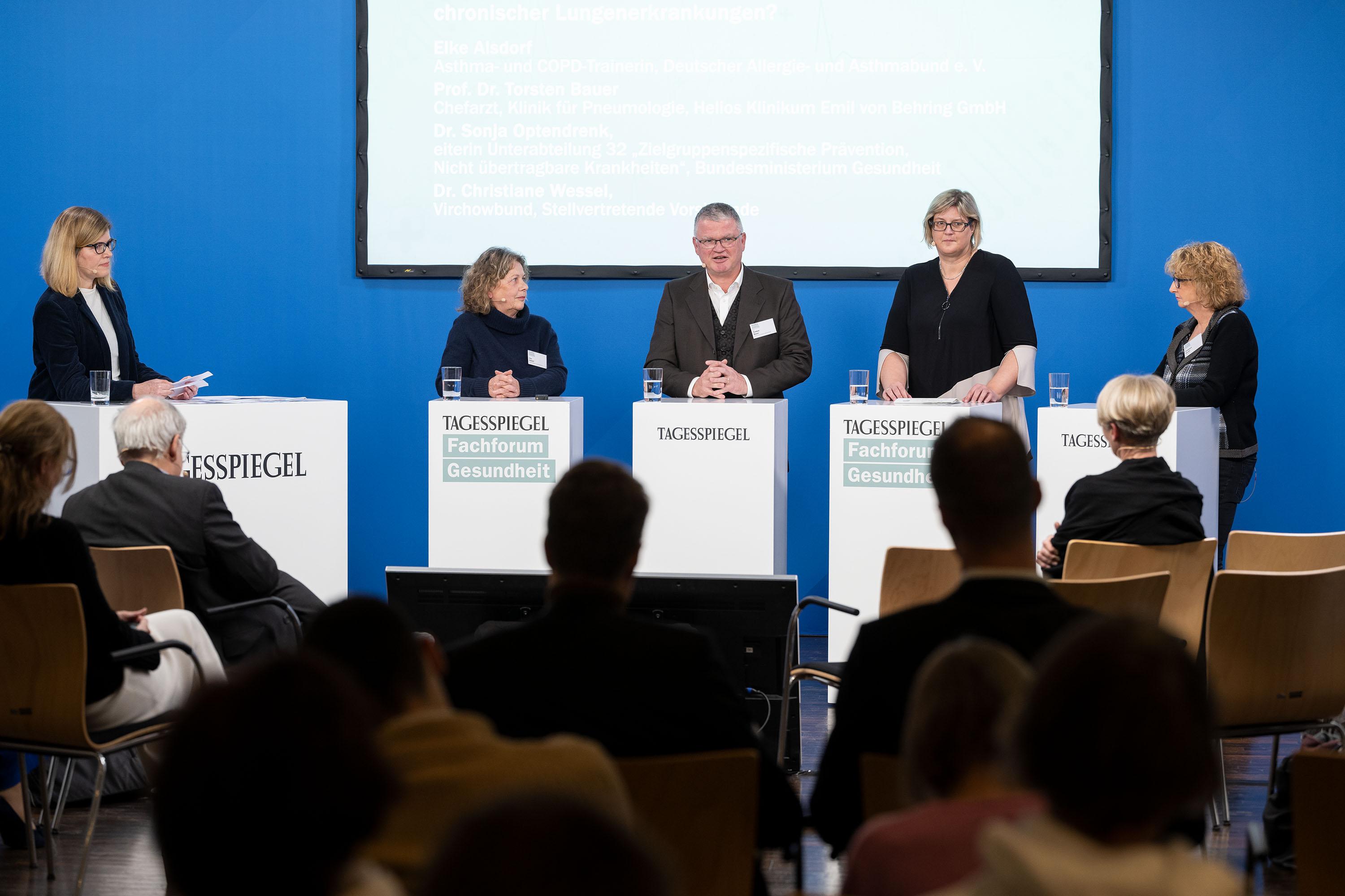 Vor Publikum stehen jeweils fünf Personen hinter einem Rednerpult. Im Hintergrund ein Screen.