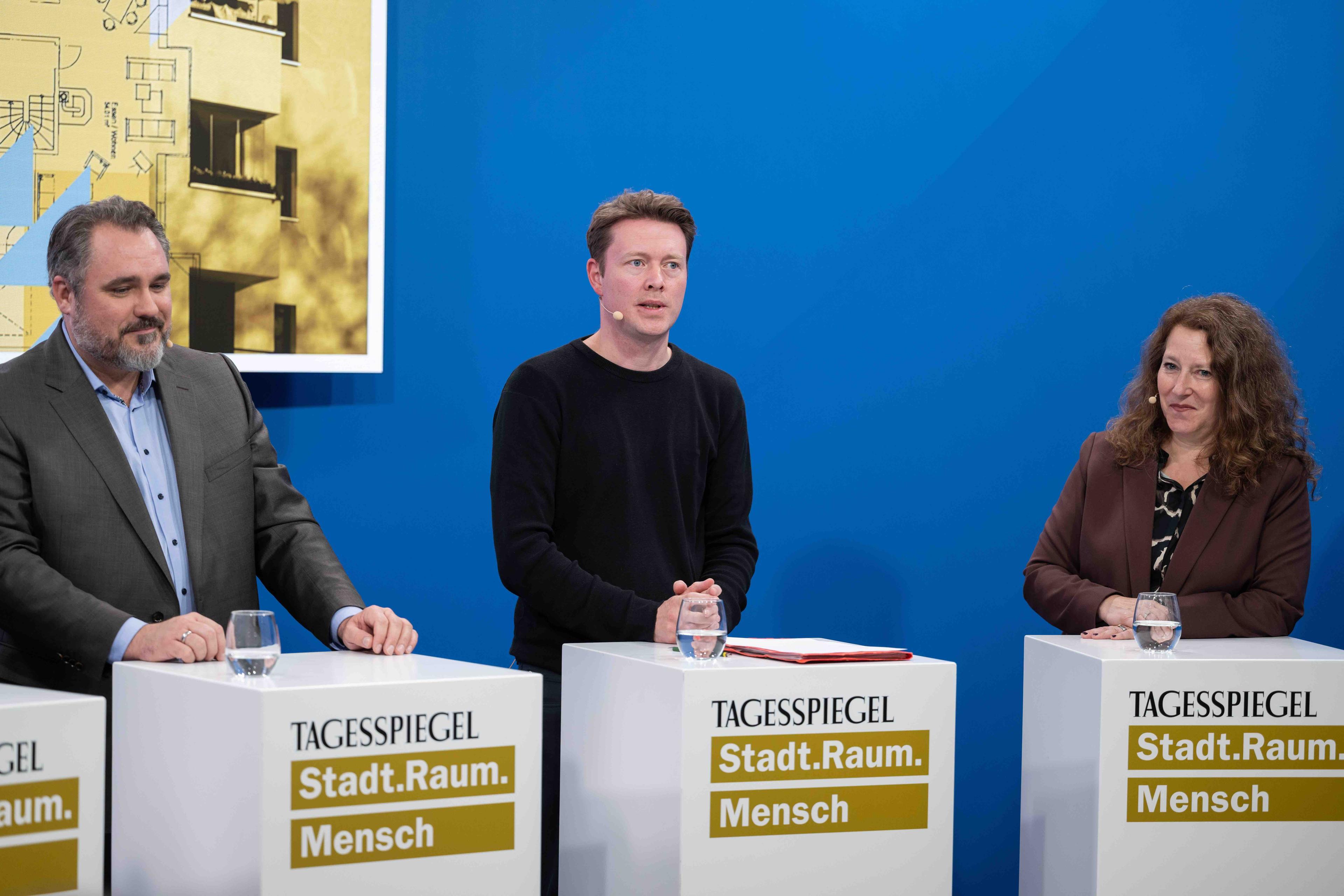 Drei Personen stehen jeweils hinter einem Rednerpult. Der Hintergrund ist Blau.
