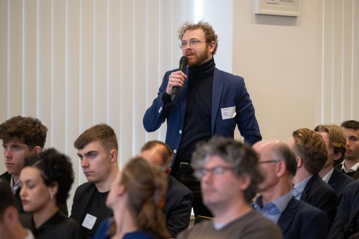 Teilnehmer steht im Publikum und stellt mit Mikrofon eine Frage; Konferenzsaal, Tageslicht.