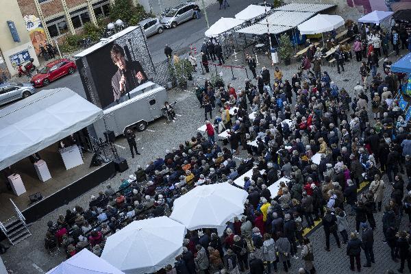 Großes Publikum bei Outdoor-Event mit Bühne, Leinwand, Zelten und weißen Sonnenschirmen.