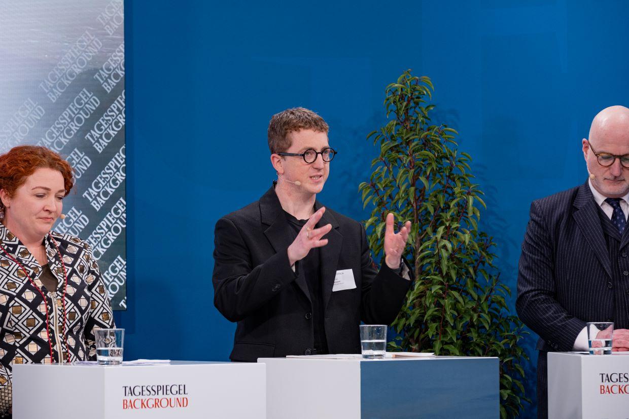 Vortragende Person spricht vor großem Bildschirm, Publikum im Vordergrund.