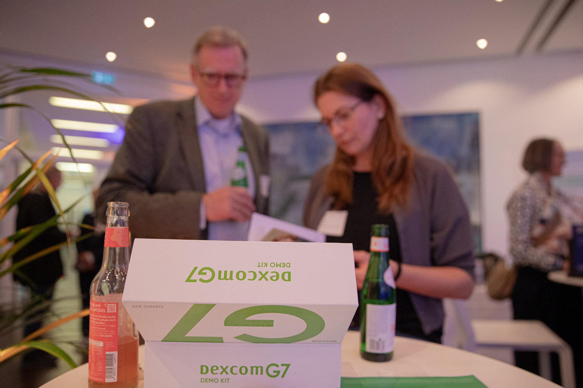 Weiße Boxen mit grüner Schrift Dexcom G7 DEMO KIT im Fokus, dahinter unscharf zwei Personen.